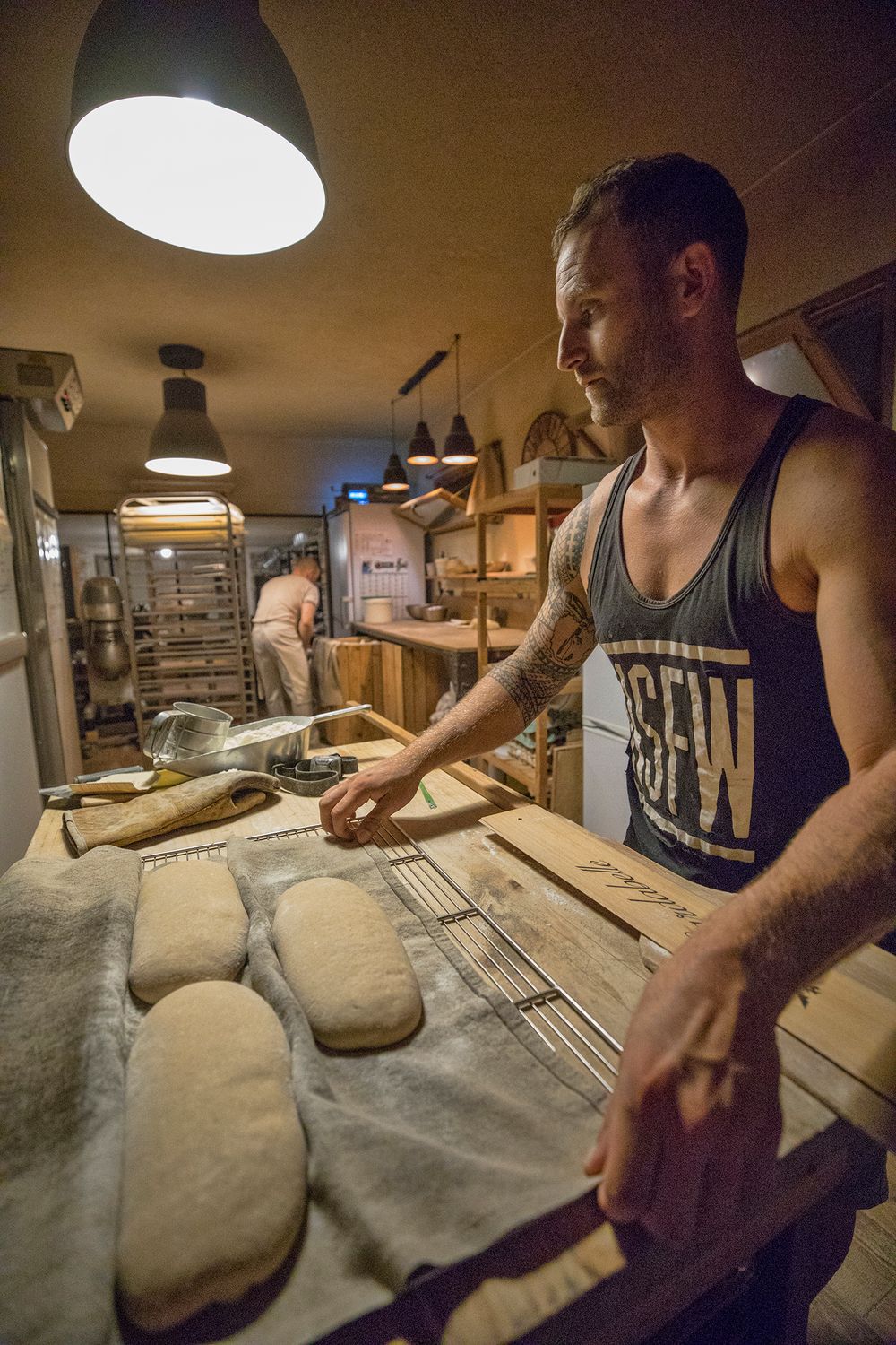 Tattooed baker arranging proofed bread loaves on linen cloth in artisan bakery.