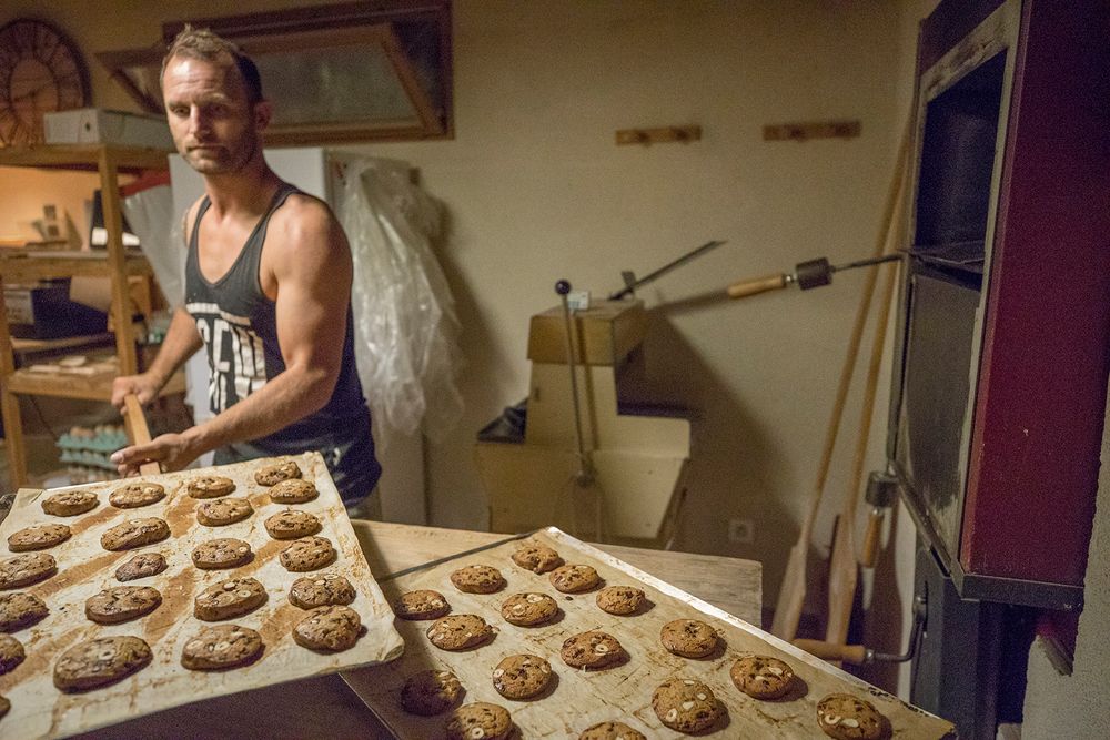 Tattooed artisan baker placing trays of freshly baked cookies near a commercial oven.