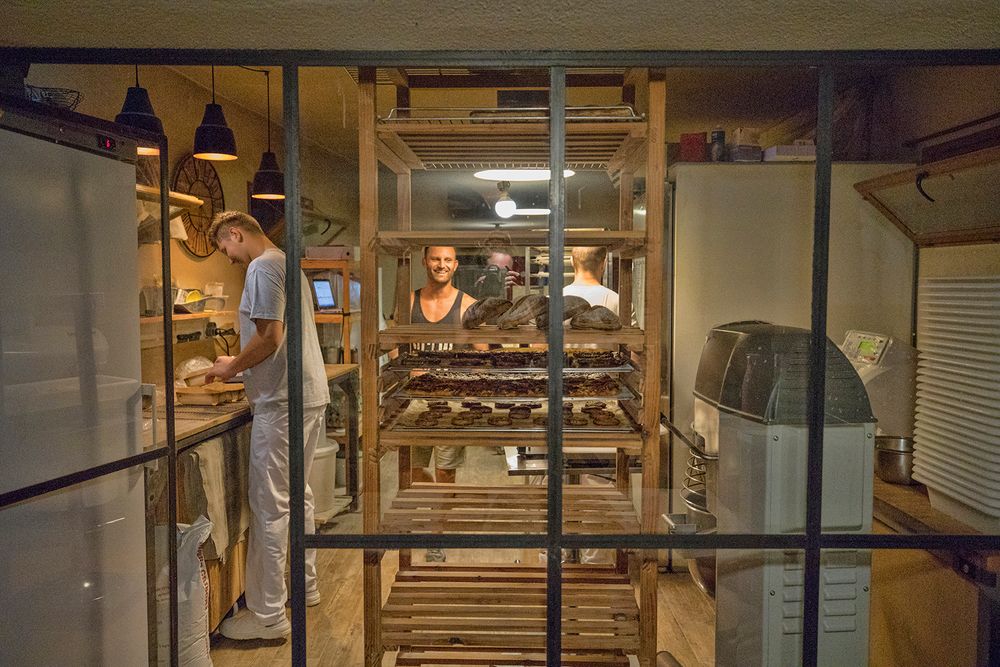 Two bakers working in a warmly lit artisan bakery kitchen viewed through glass partition with bread racks.