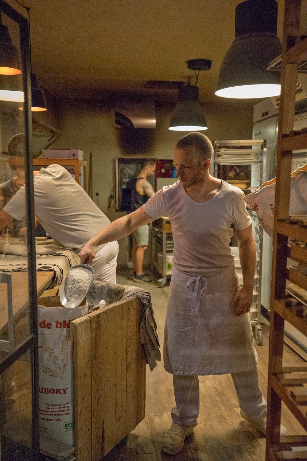 Male baker in white apron pouring flour into a mixing bin in a professional artisan bakery kitchen.