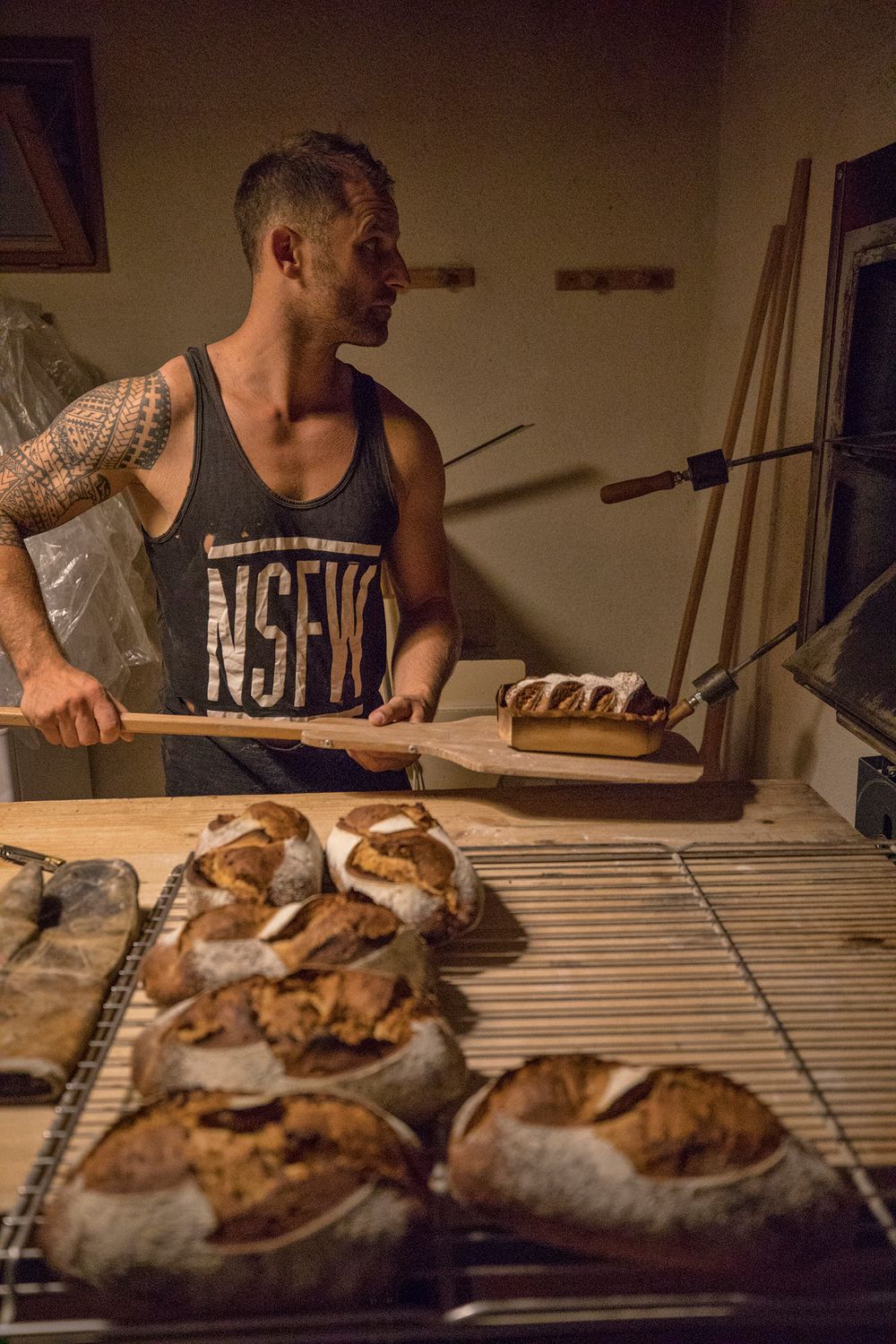 Tattooed male baker using a wooden peel to load bread into a stone oven with rustic sourdough loaves.