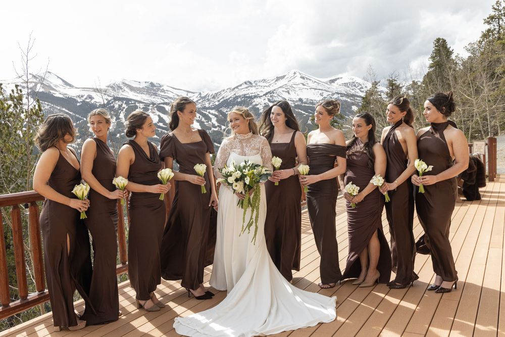 Bride in white gown with bridesmaids in brown dresses on mountain deck with snowy peaks backdrop.
