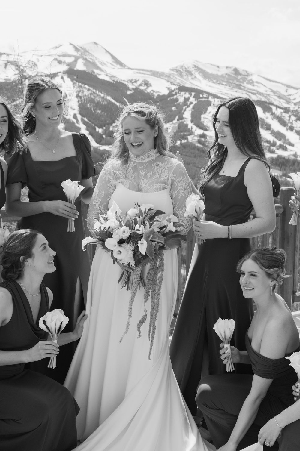 Bride in lace gown holds bouquet surrounded by bridesmaids in black dresses with snowy mountain backdrop.