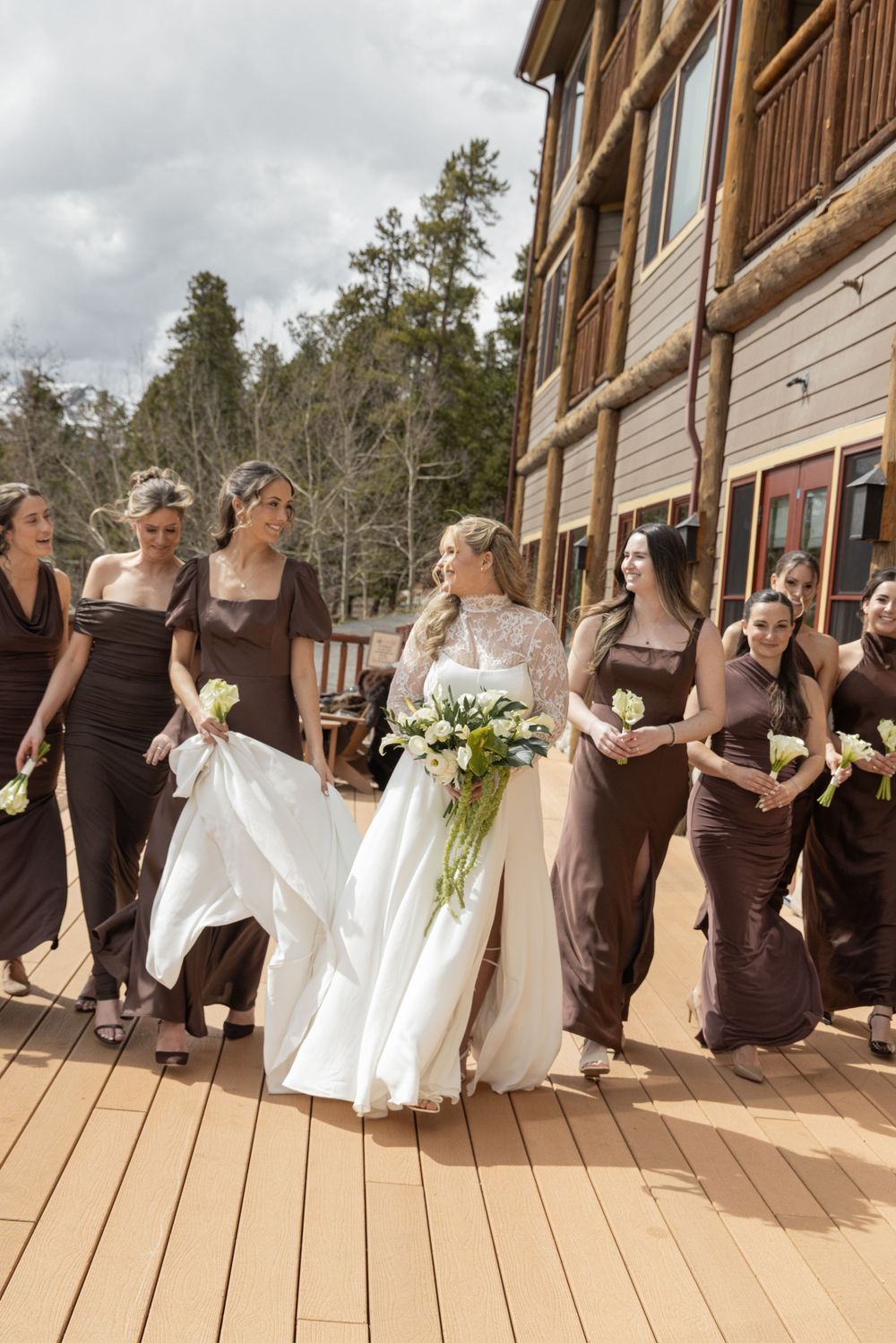 Bride in white gown walks with bridesmaids in brown dresses on wooden deck outside rustic log cabin venue.