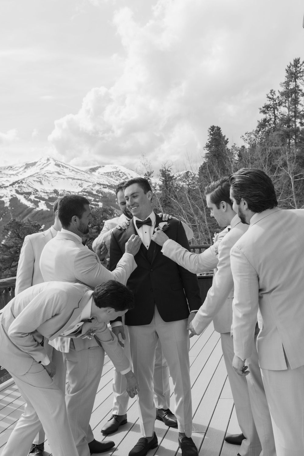 Groomsmen help groom adjust boutonniere on deck with snowy mountain backdrop in black and white wedding photo.