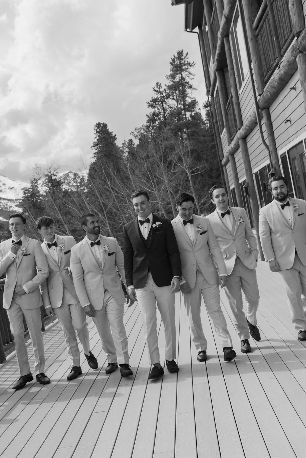 Groom and groomsmen in light suits and bow ties walk together on a deck in a black and white wedding photo.