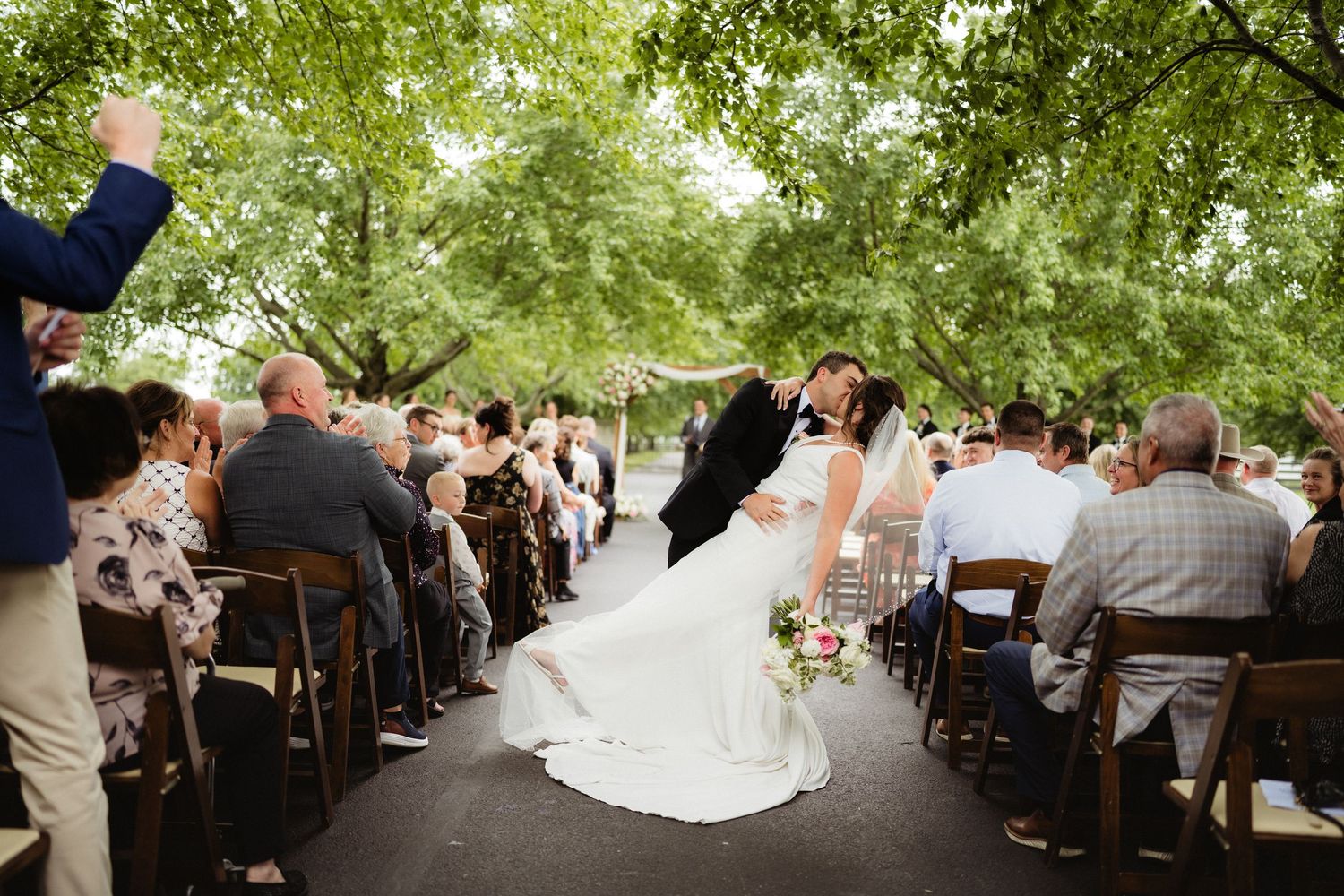 Bride and groom kiss while walking down the aisle at an outdoor wedding ceremony under lush green trees.