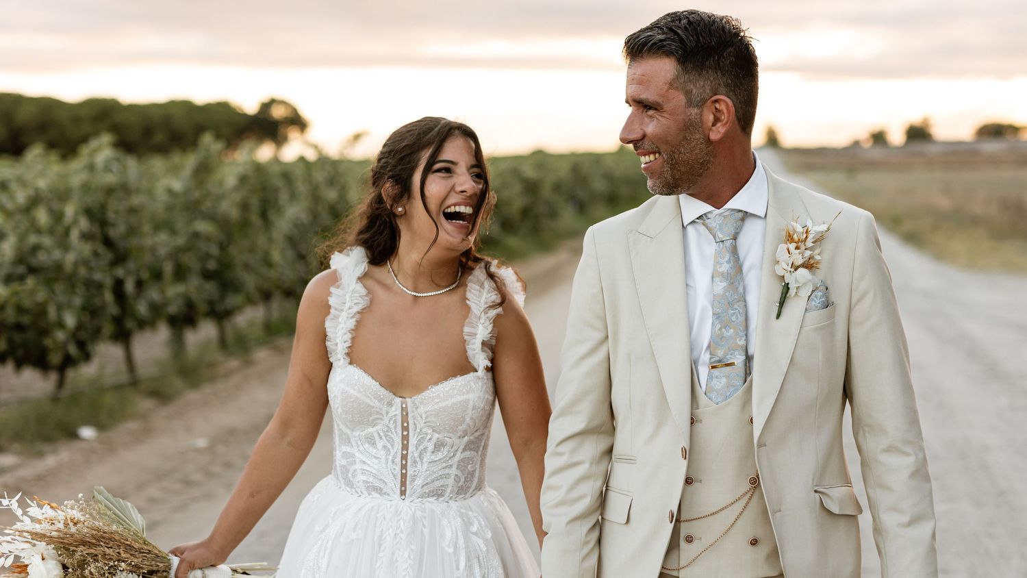 A joyful bride and groom share a candid moment on a vineyard path during sunset.
