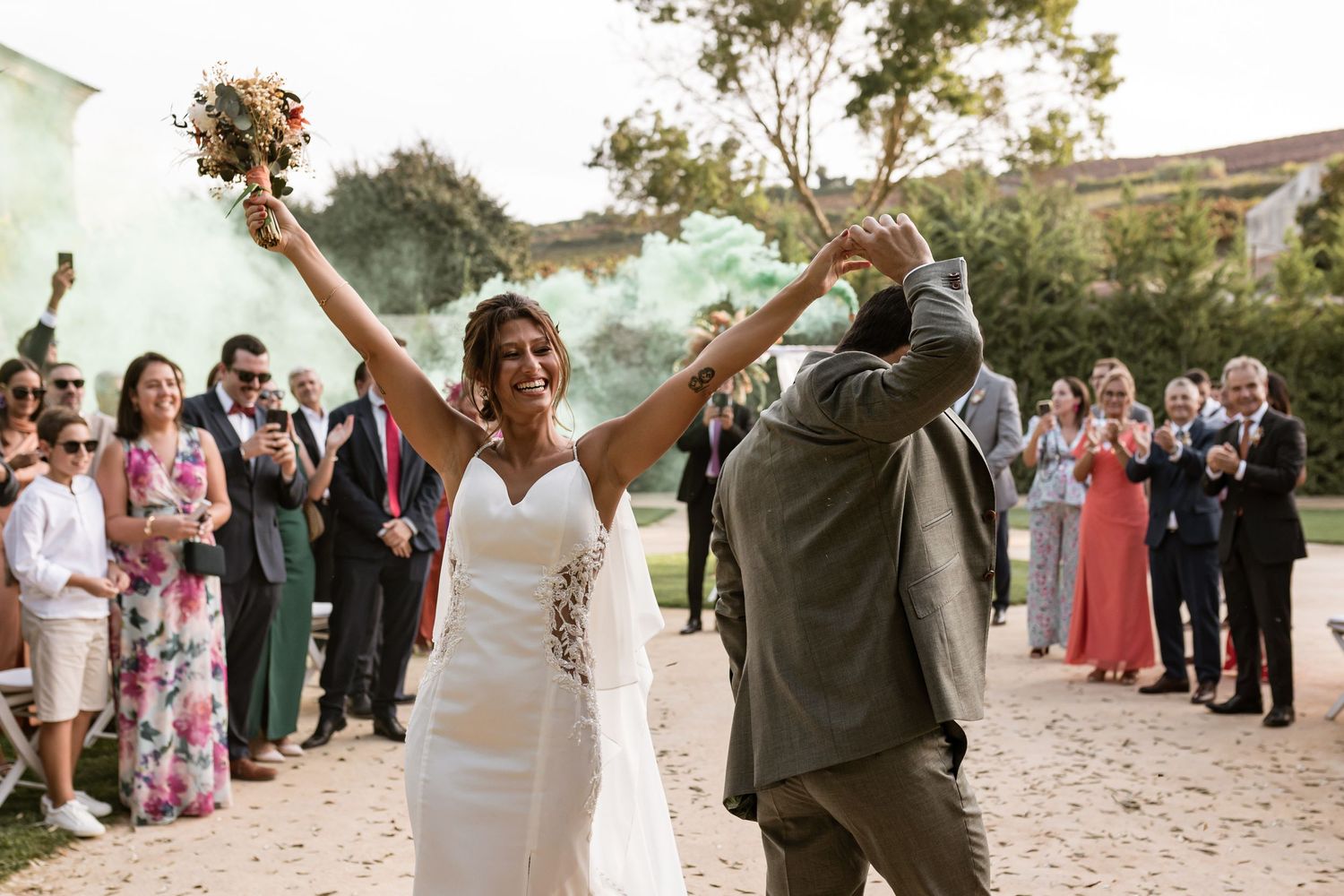 Newlyweds celebrate with raised arms surrounded by wedding guests applauding on a sunny outdoor path.