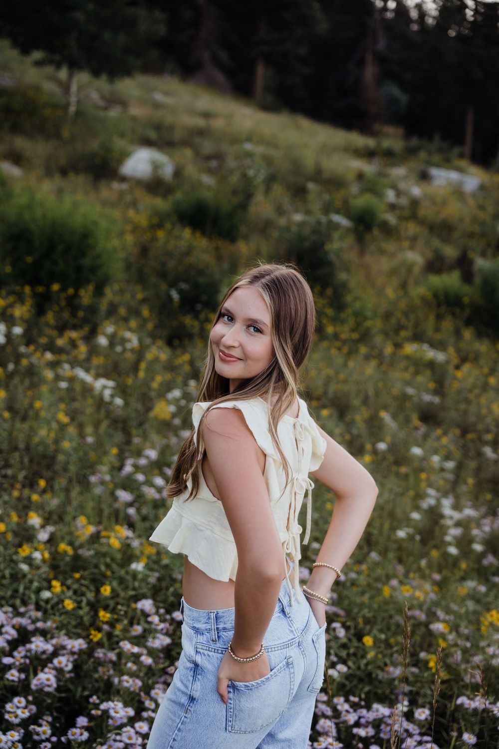 Young woman in white crop top and jeans smiling in a wildflower meadow.