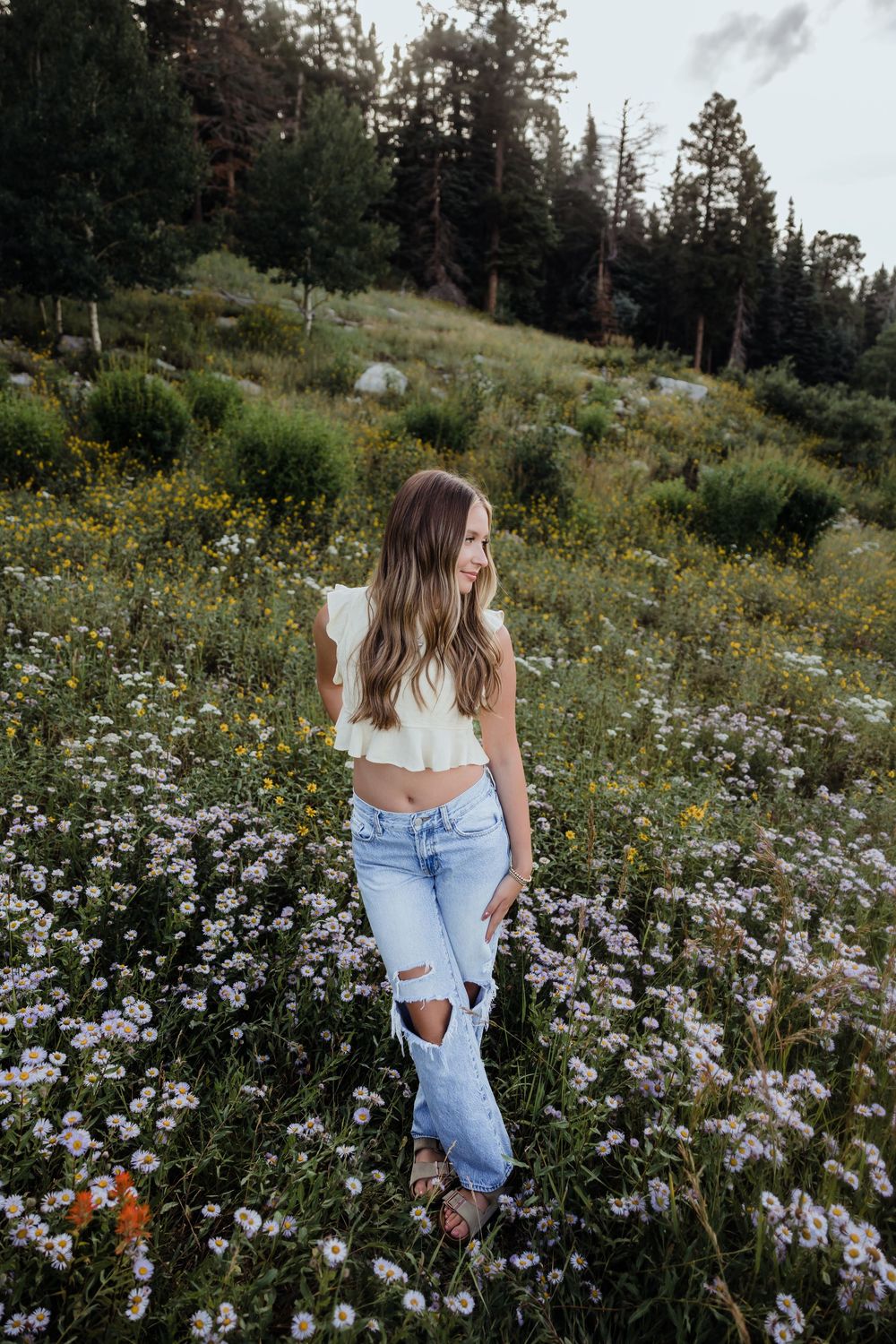 Young woman in white crop top and ripped jeans standing in a wildflower meadow surrounded by pine trees.