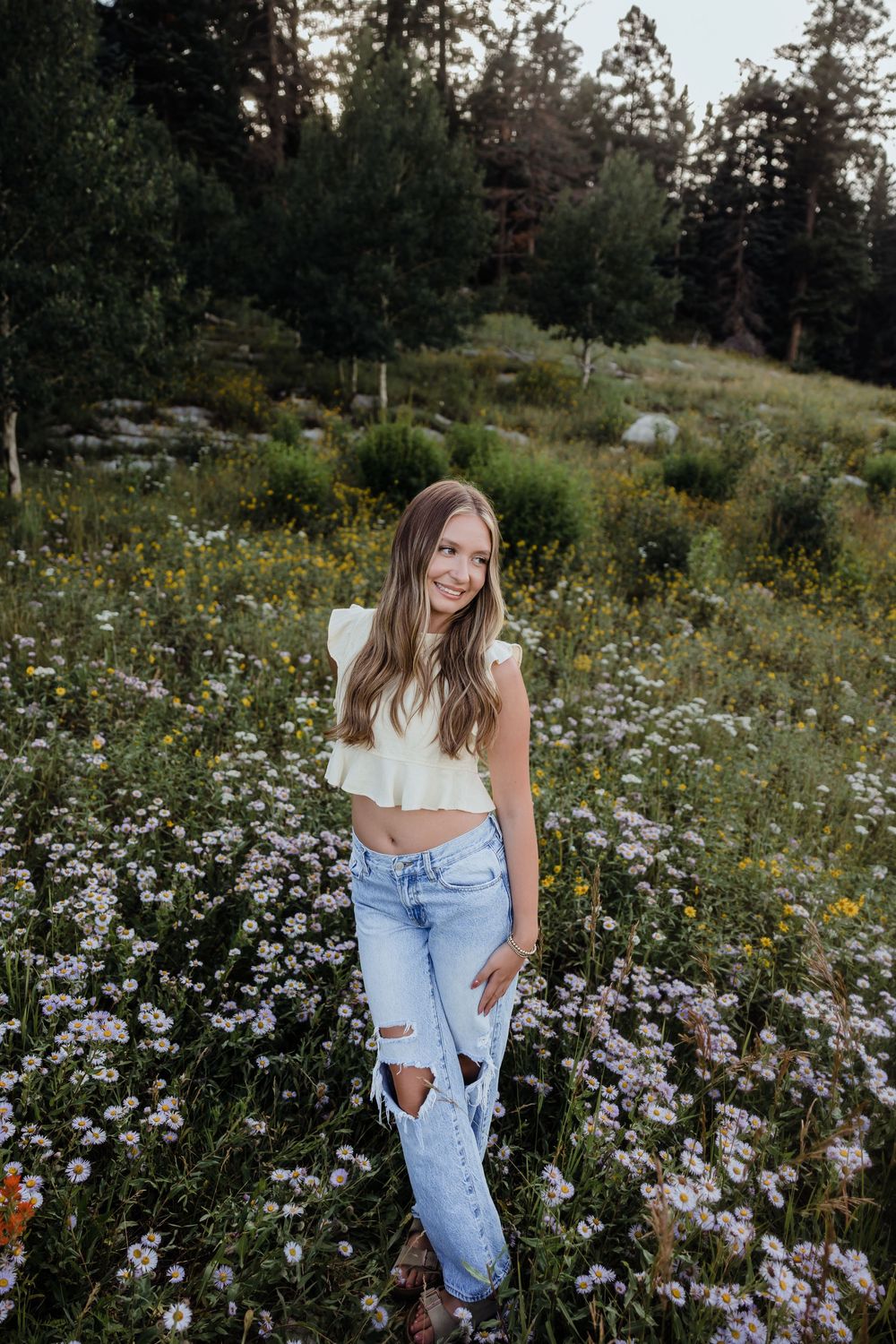 Young woman in white crop top and ripped jeans smiling in a wildflower meadow surrounded by pine trees.