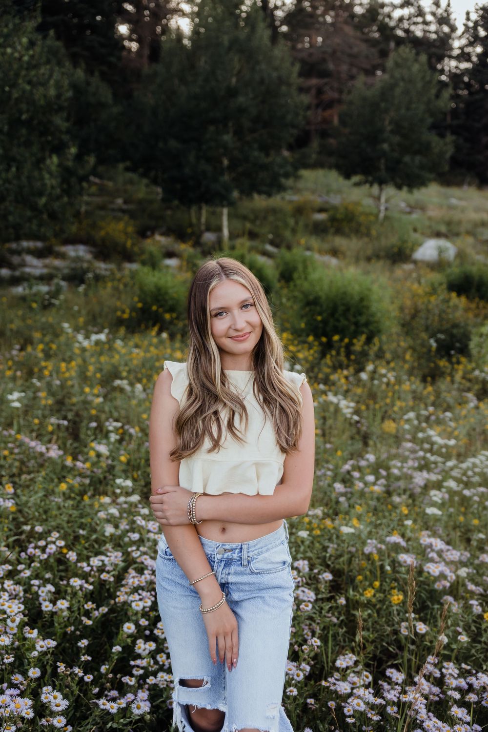 Young woman in white crop top and ripped jeans posing in a wildflower meadow with trees in background.