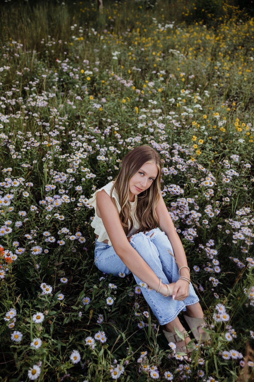 Young woman sitting in a wildflower meadow with daisies, wearing jeans and a light top, looking at camera.