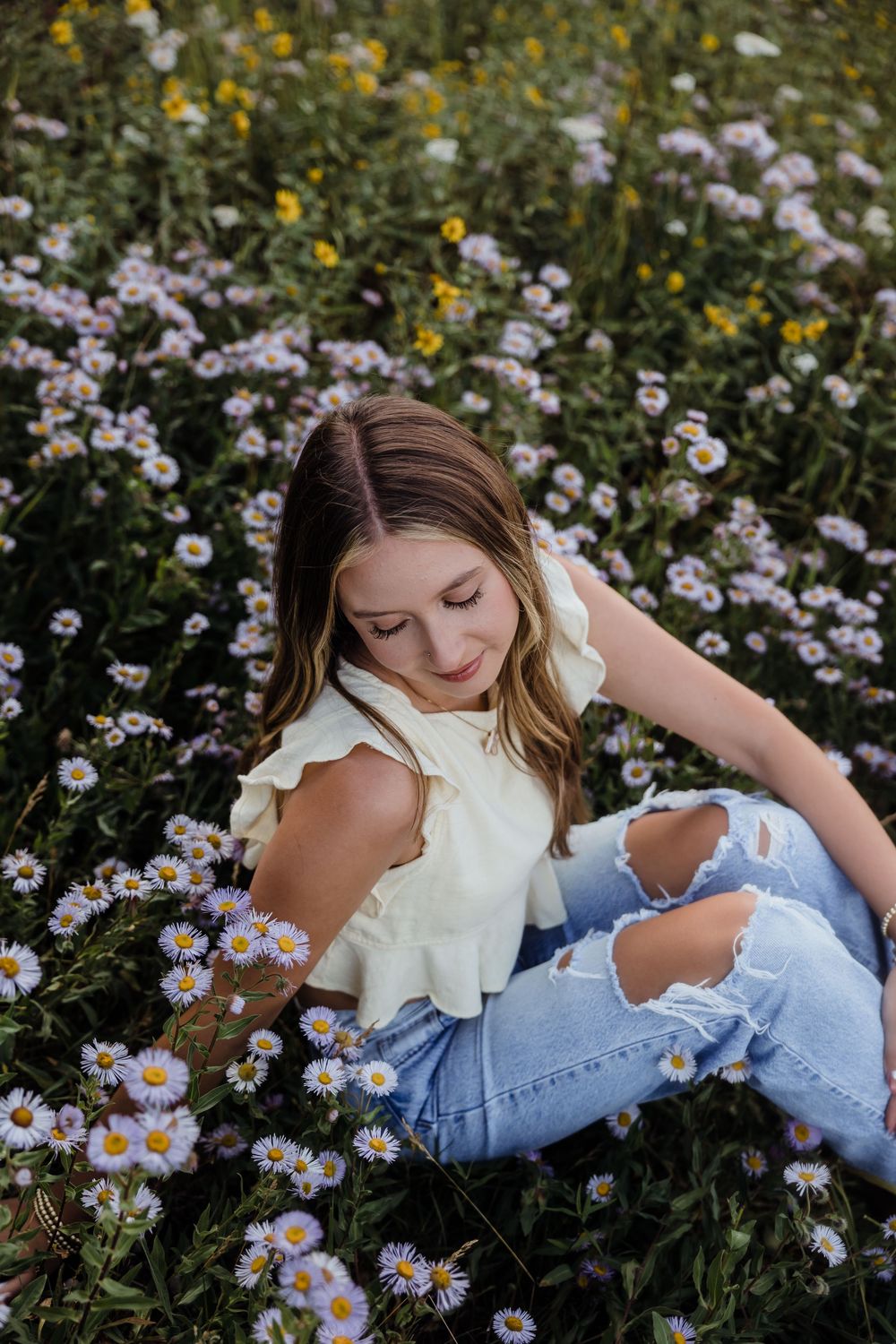 Young woman in white ruffled top and ripped jeans sitting among blooming wildflowers and daisies in a lush garden.