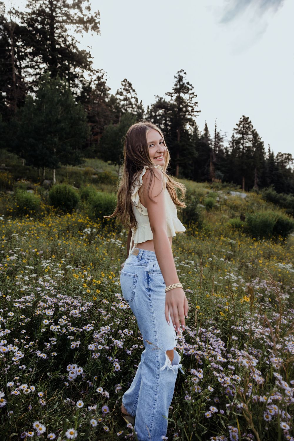 Young woman in crop top and jeans smiling in a wildflower meadow surrounded by pine trees.