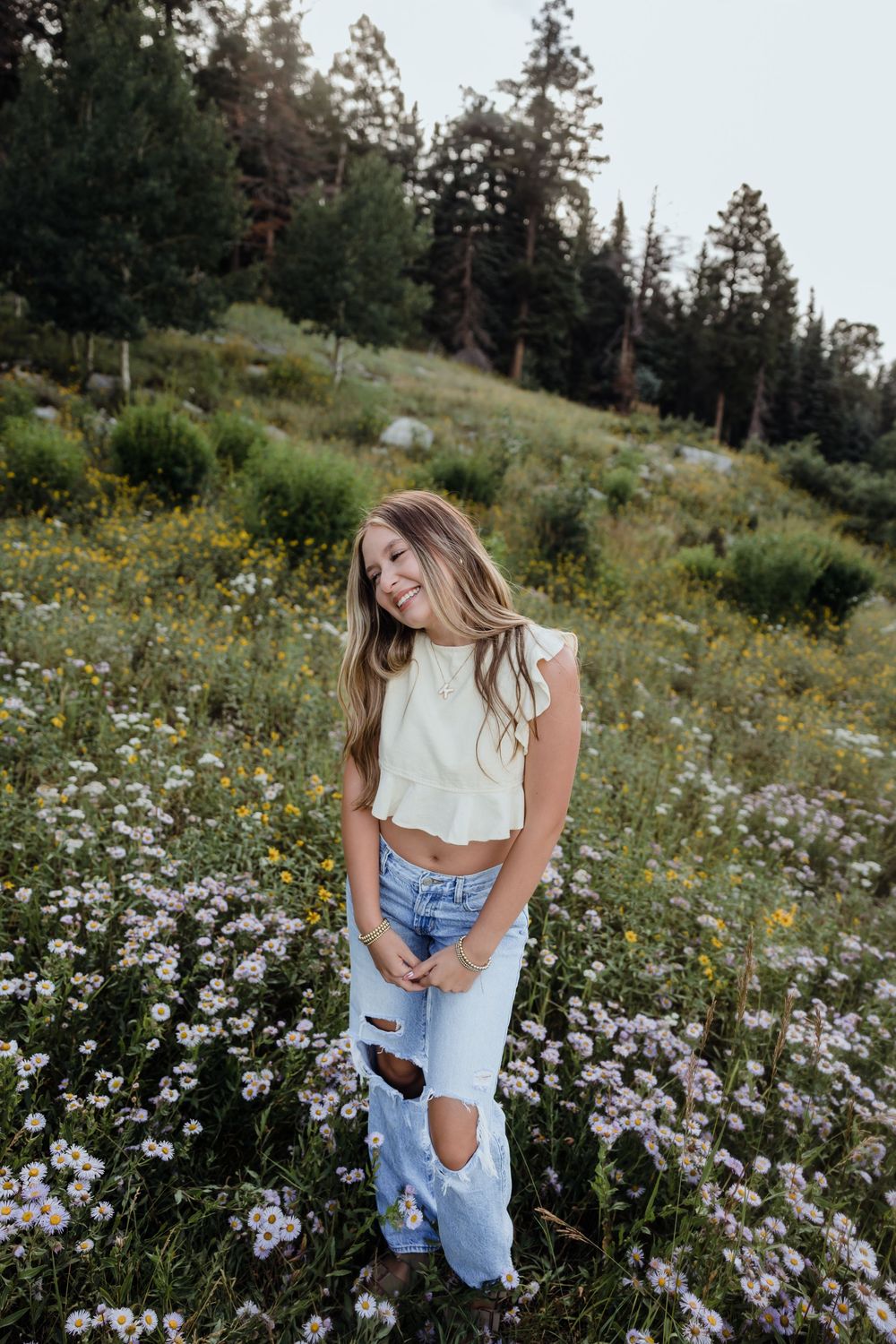 Smiling young woman in white crop top and ripped jeans standing in a wildflower meadow surrounded by pine trees.