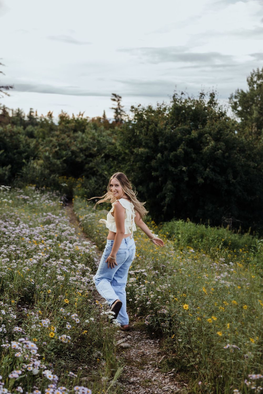 Smiling young woman in white crop top and blue jeans walking through a wildflower field path.