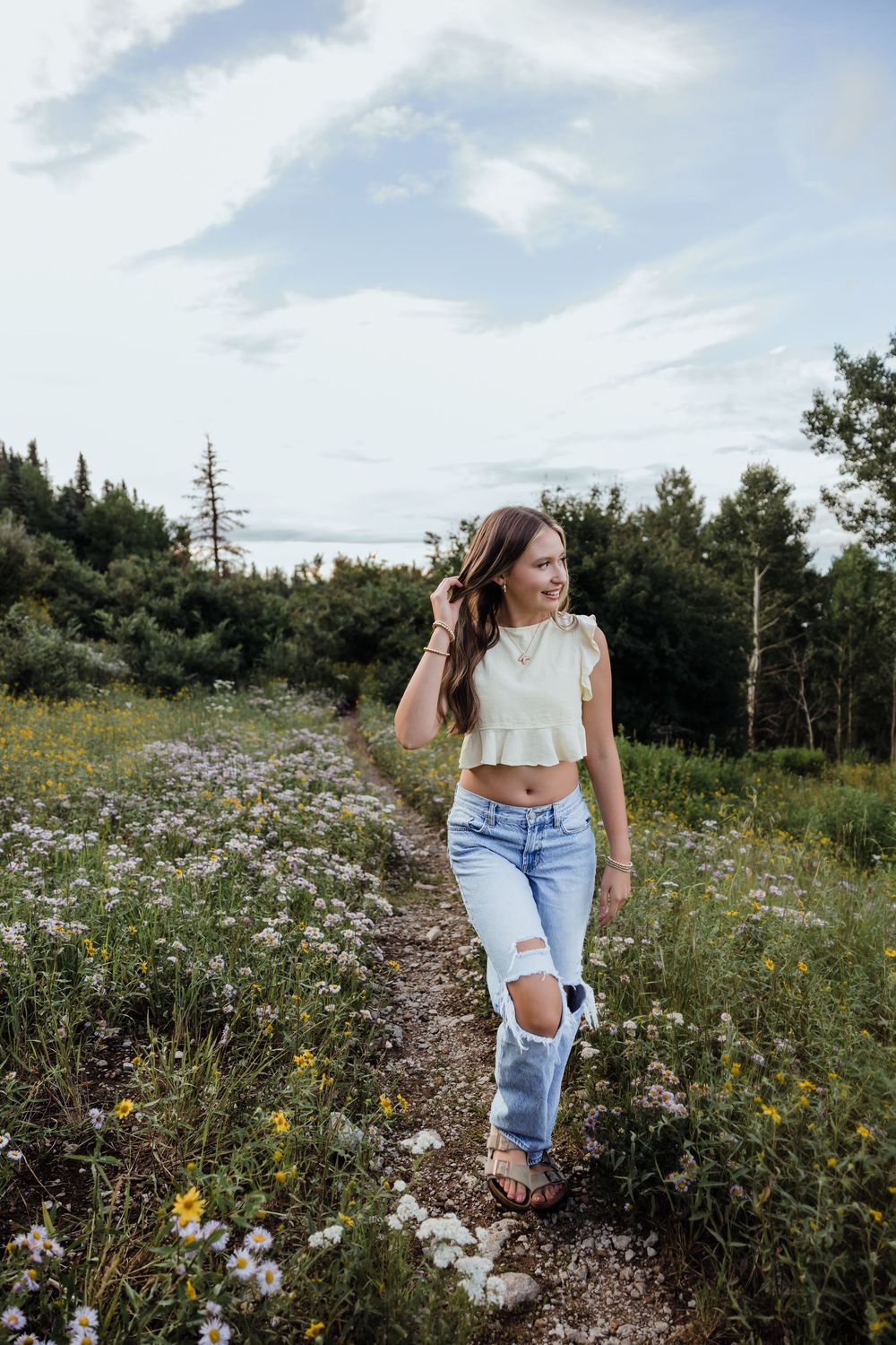Young woman in white crop top and ripped jeans walking along a wildflower meadow path with trees in background.