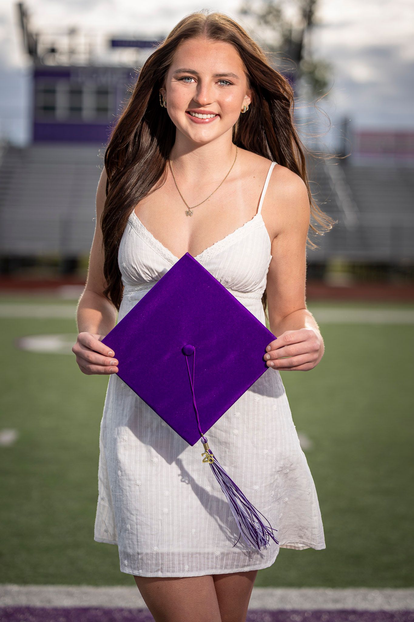 Smiling young woman in white dress holding purple graduation cap on football field.