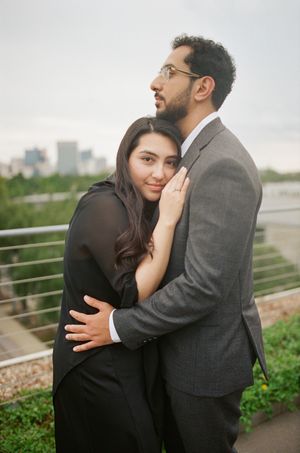 A couple embraces tenderly outdoors, woman in black outfit leaning on man in gray suit, city skyline in background.