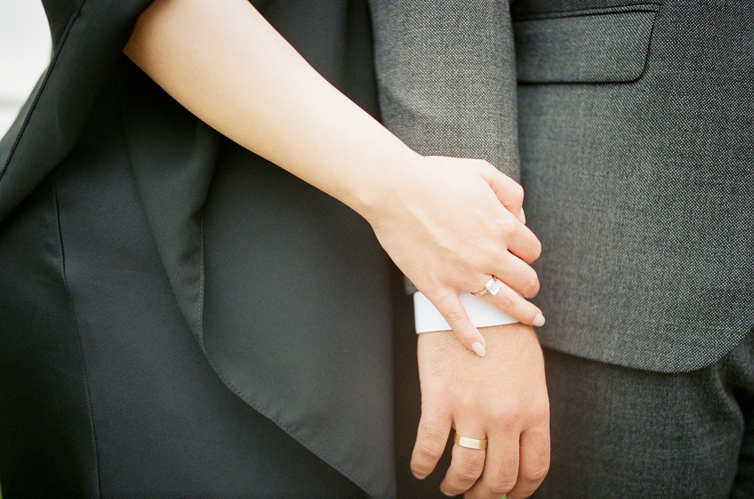 Close-up of a couple holding hands, wearing wedding rings, dressed in formal black and gray attire.