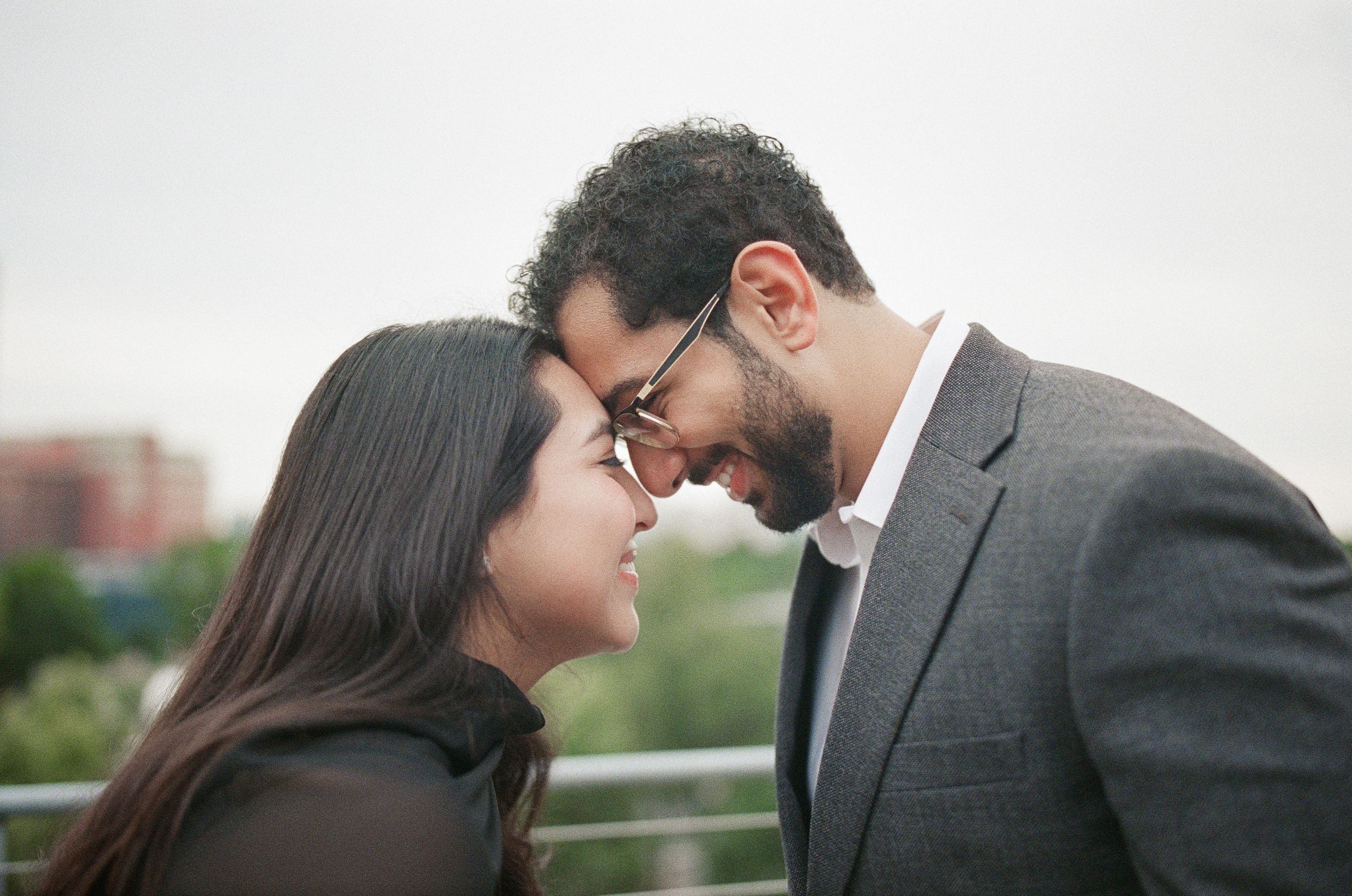 A romantic couple sharing an intimate moment, touching foreheads outdoors, man wearing glasses and gray suit.