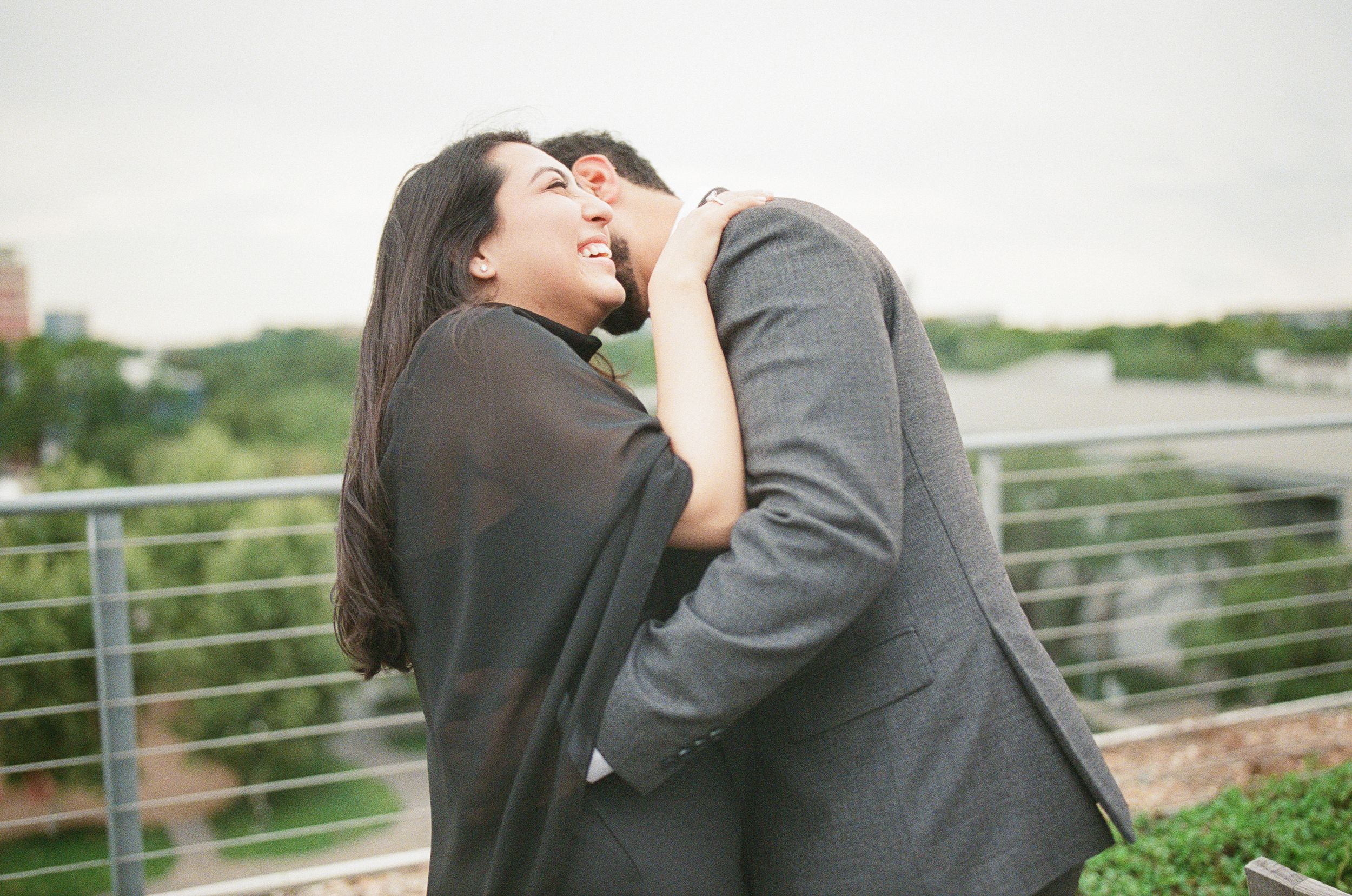 Couple sharing a romantic embrace and laughing on an outdoor rooftop terrace with city views.