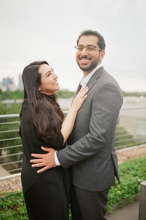 A smiling couple in formal attire embraces outdoors, woman in black dress, man in gray suit with glasses.