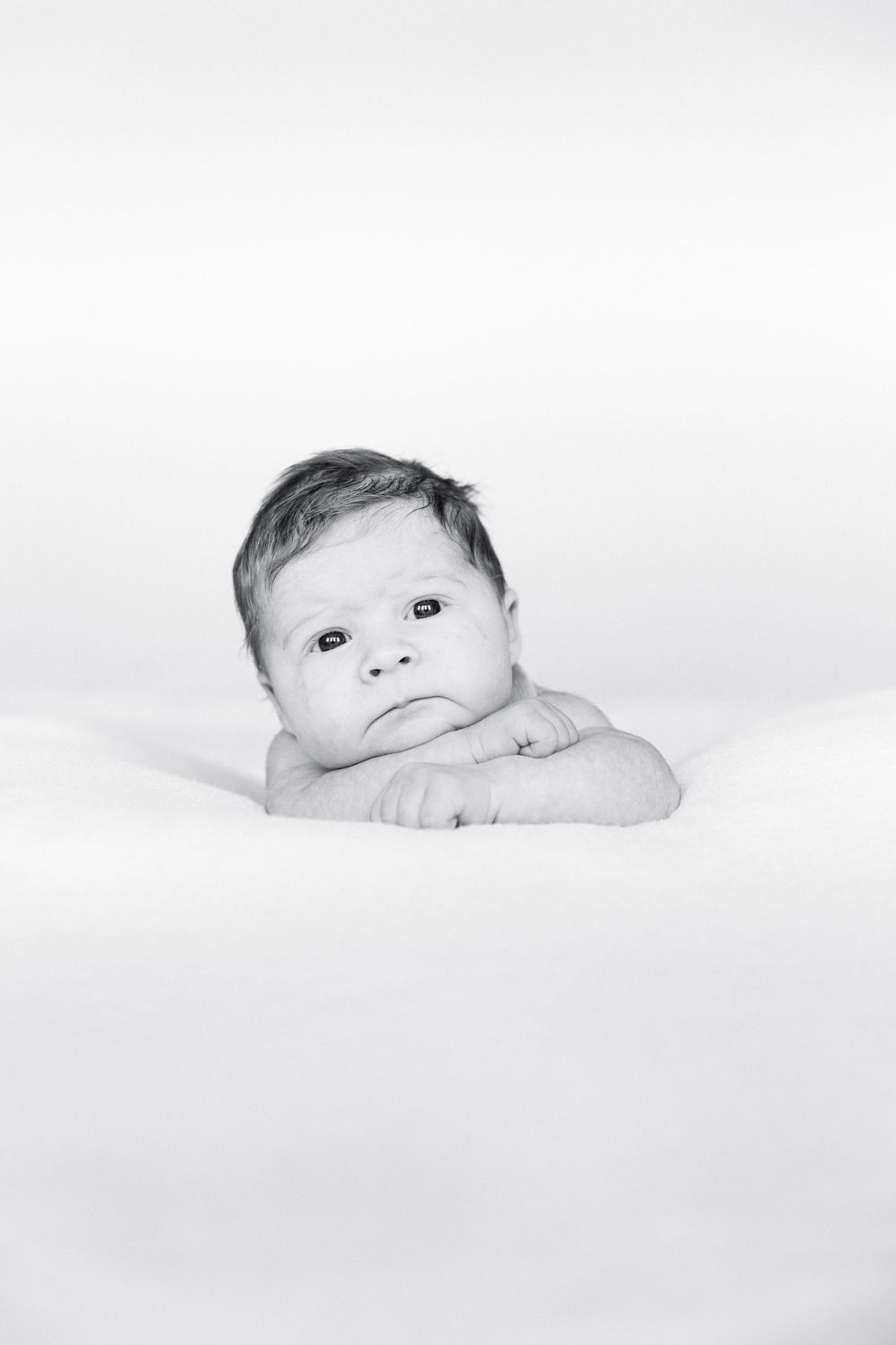 Black and white portrait of an alert wide-eyed newborn baby resting chin on folded hands.