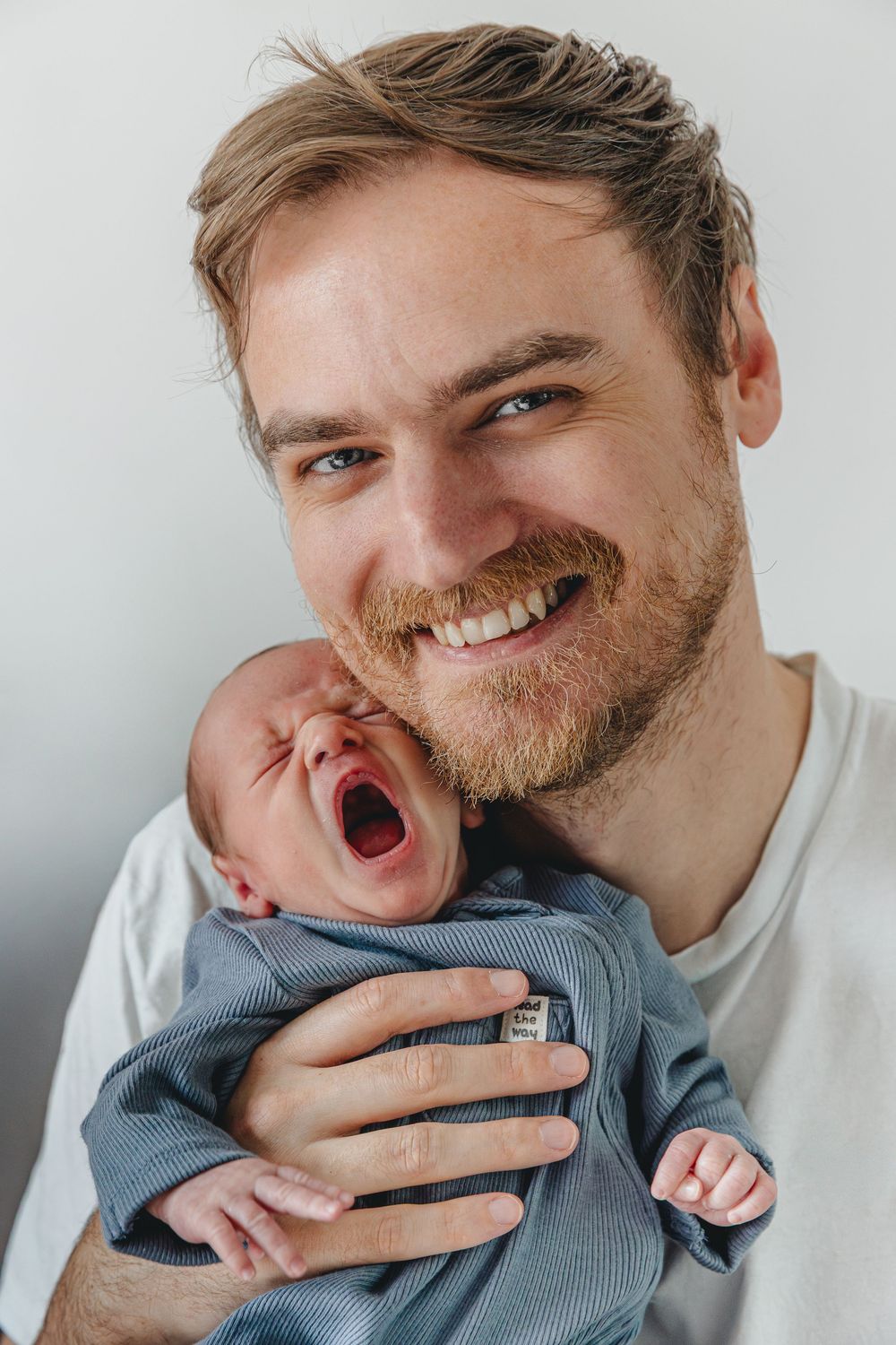 Smiling bearded father holding yawning newborn baby against a light gray background.