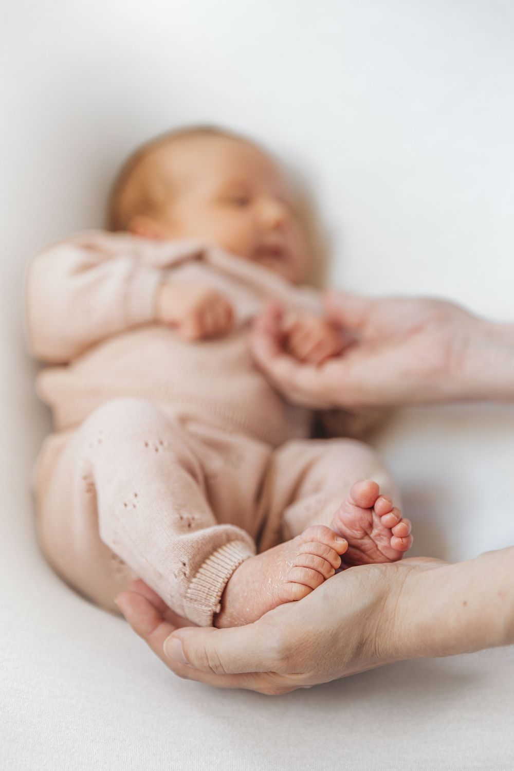 Newborn baby in pink outfit lying on white surface with parent's hands holding tiny feet.