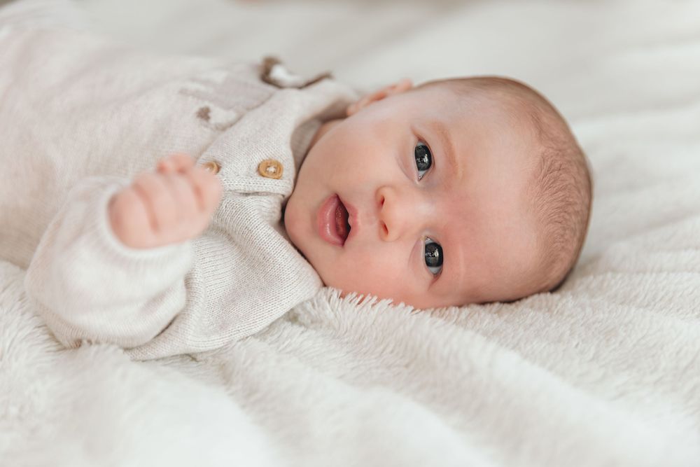 Cute wide-eyed newborn baby in white onesie lying on soft white blanket.