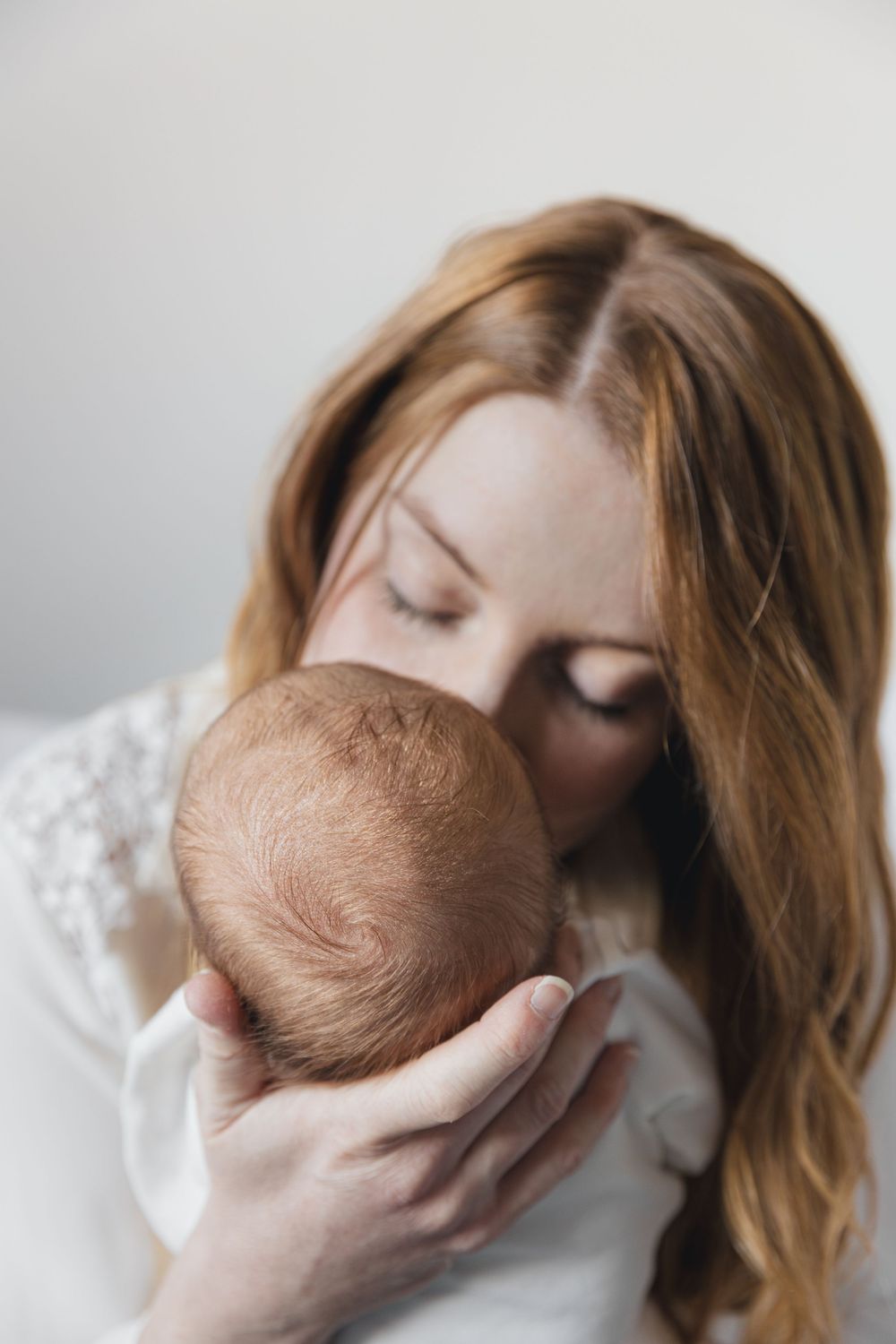 Loving red-haired mother gently kissing her newborn baby's head against white background.