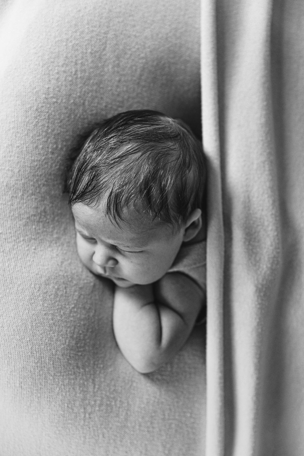 Black and white photo of a sleeping newborn baby nestled in soft draped fabric.