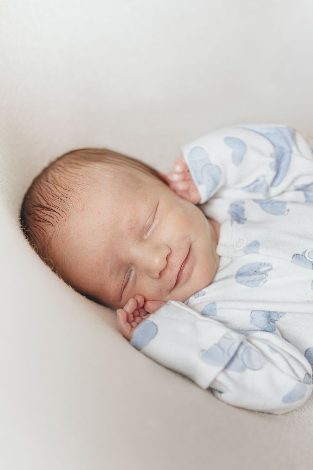 Peaceful newborn baby sleeping in blue and white patterned onesie with hands tucked under cheek.
