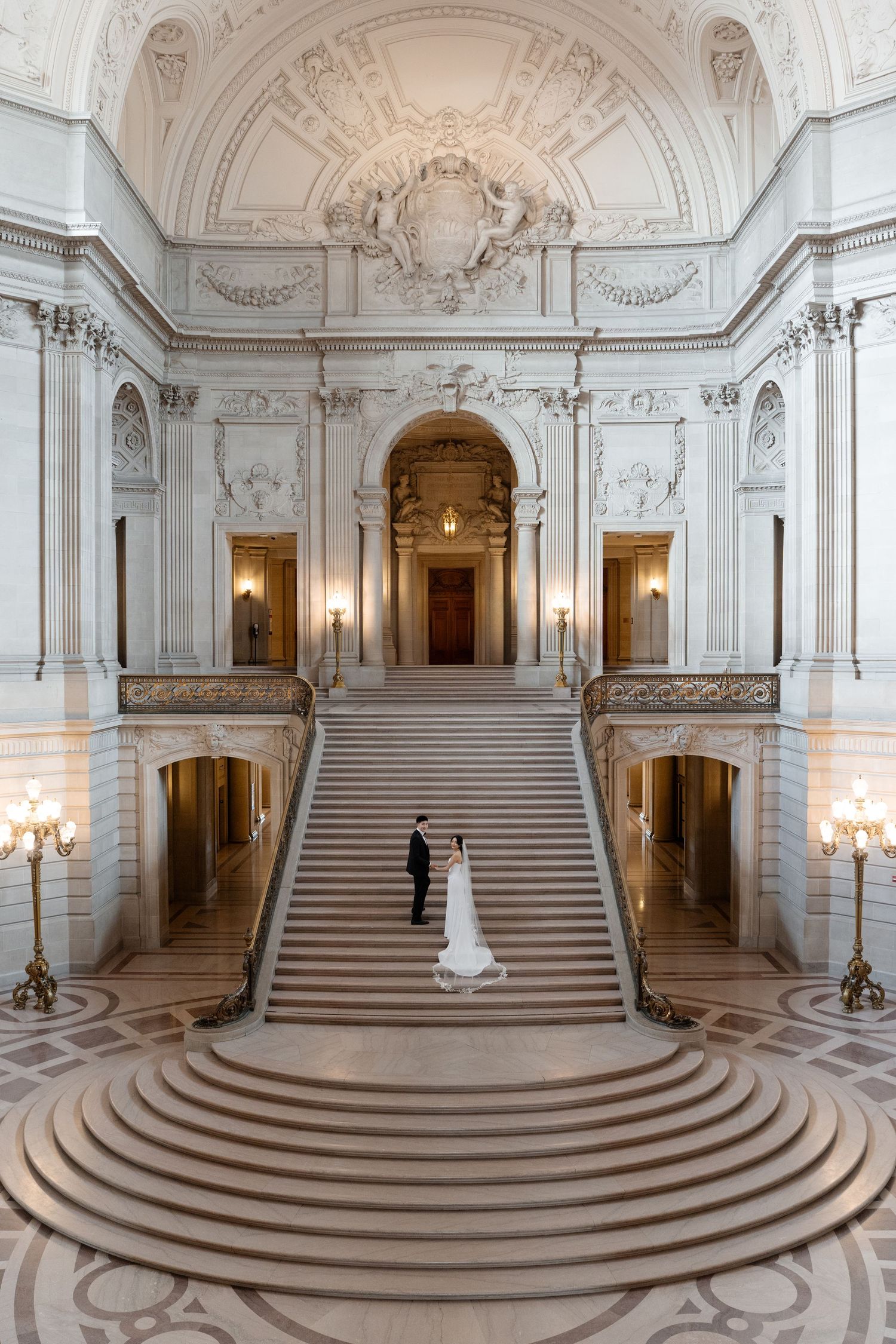 Bride and groom embrace on the grand staircase of San Francisco City Hall, surrounded by ornate Beaux-Arts architecture.