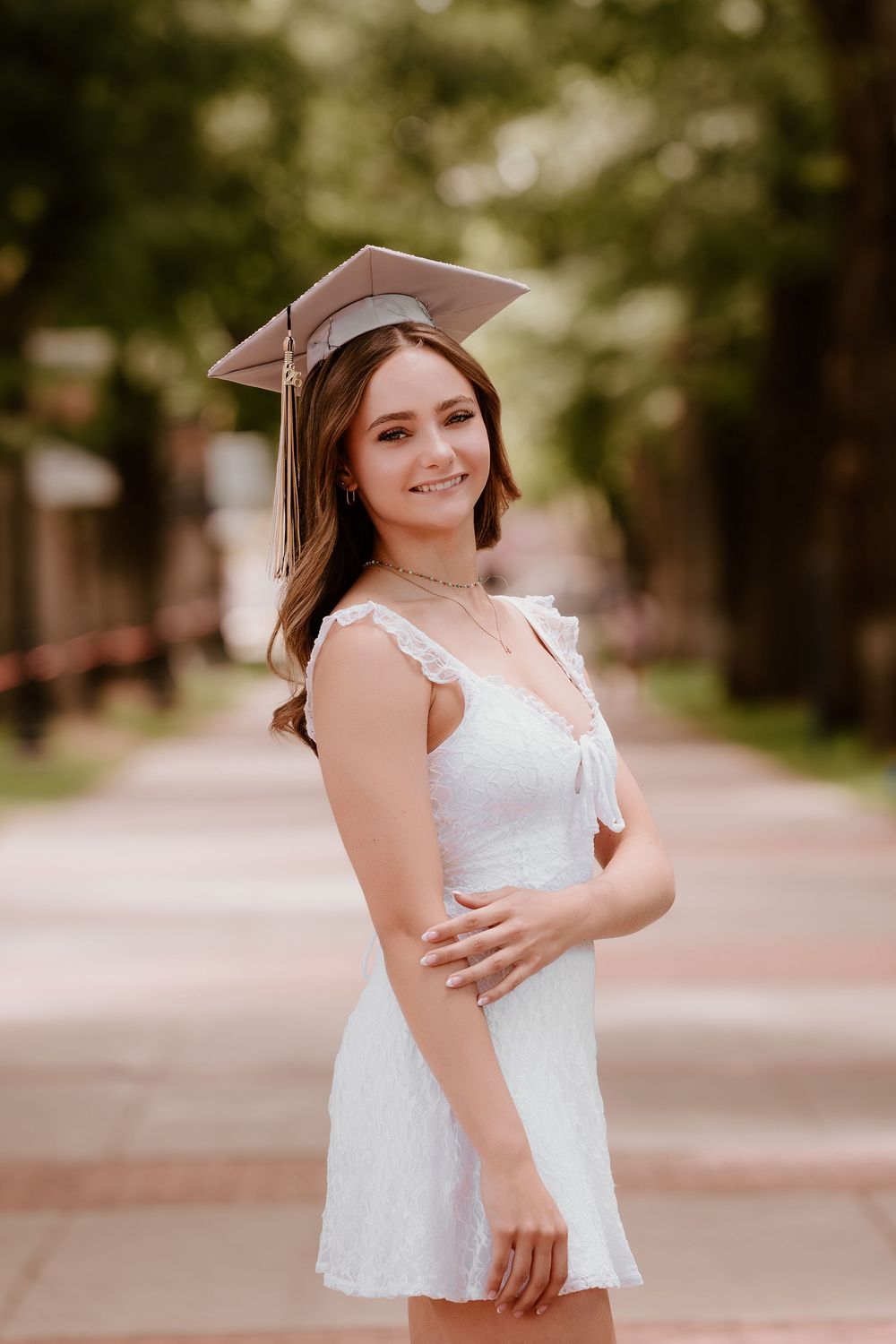 Young woman in white dress and graduation cap smiling outdoors on a tree-lined campus walkway.