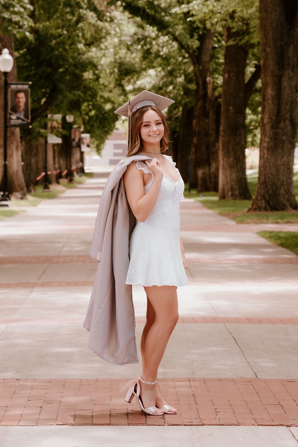 Young woman in white dress and graduation cap holding gown over shoulder on tree-lined campus walkway.