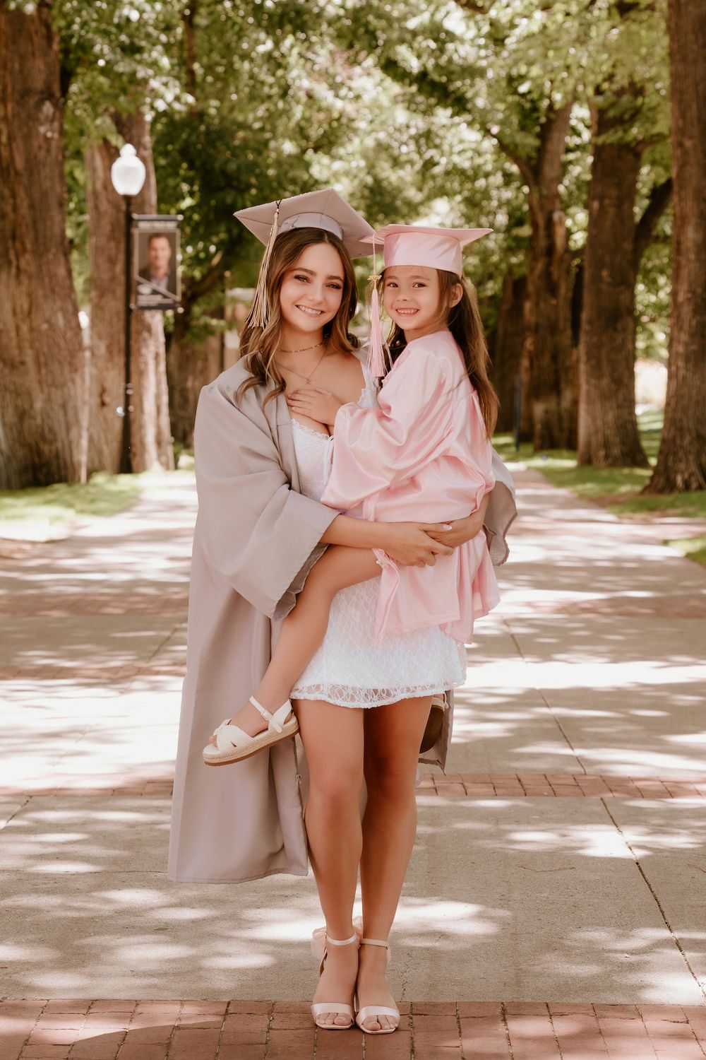 Two young women in graduation caps and gowns pose together outdoors on a tree-lined brick walkway.