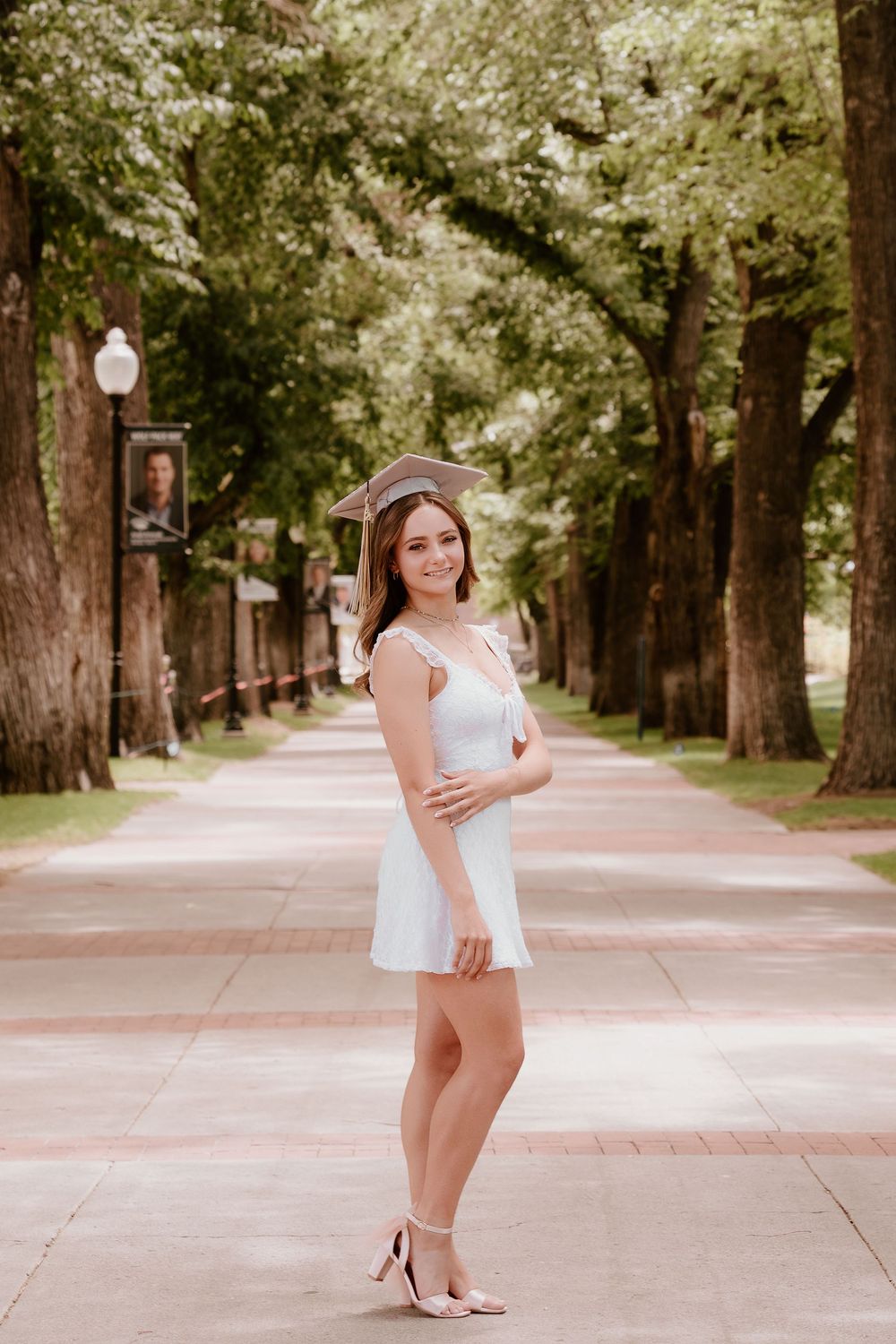Young woman in white dress and graduation cap poses on a tree-lined campus walkway celebrating her graduation.