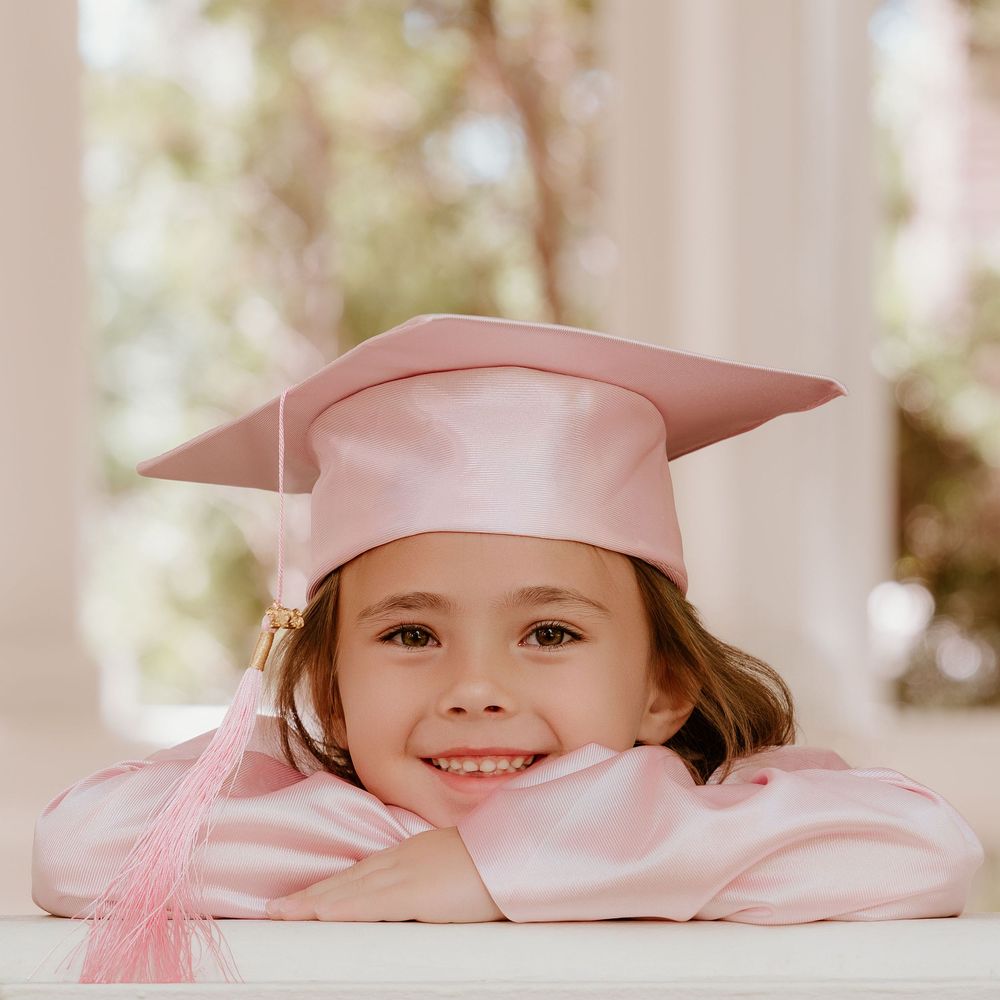 A smiling young girl wearing a pink graduation cap and gown, resting her chin on her arms at a table.