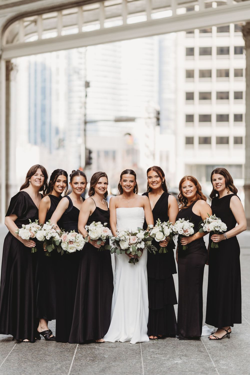 Bride in white gown poses with six bridesmaids in black dresses holding white floral bouquets.