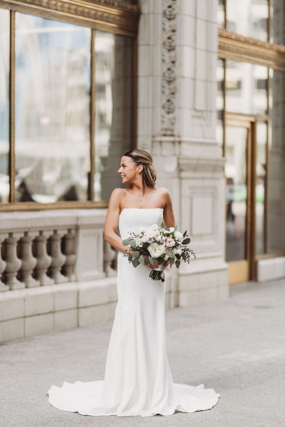Smiling bride in strapless white gown holding floral bouquet stands on ornate stone balcony of elegant building.