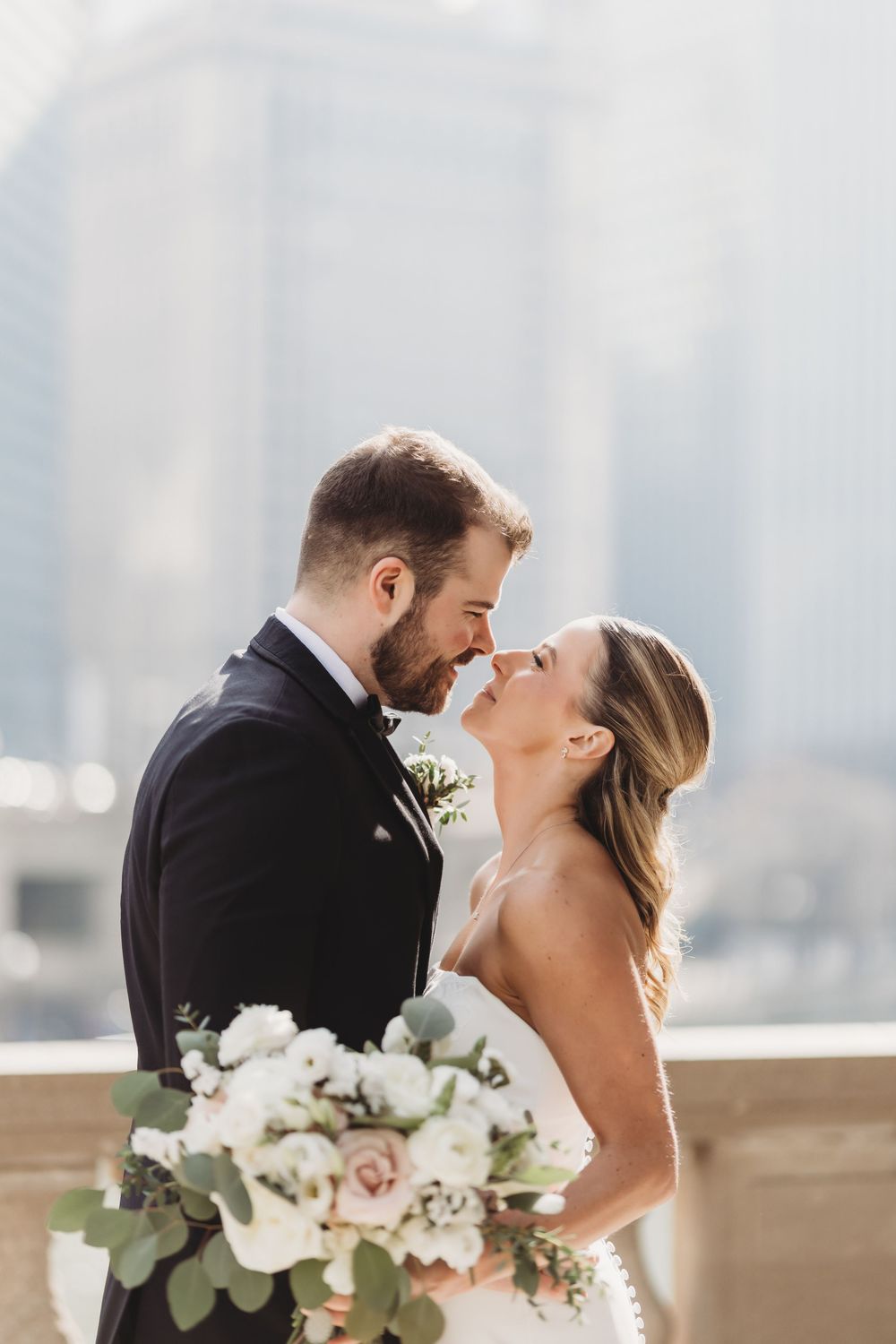 Bride and groom sharing a romantic moment, nearly kissing, with a white floral bouquet and city skyline backdrop.