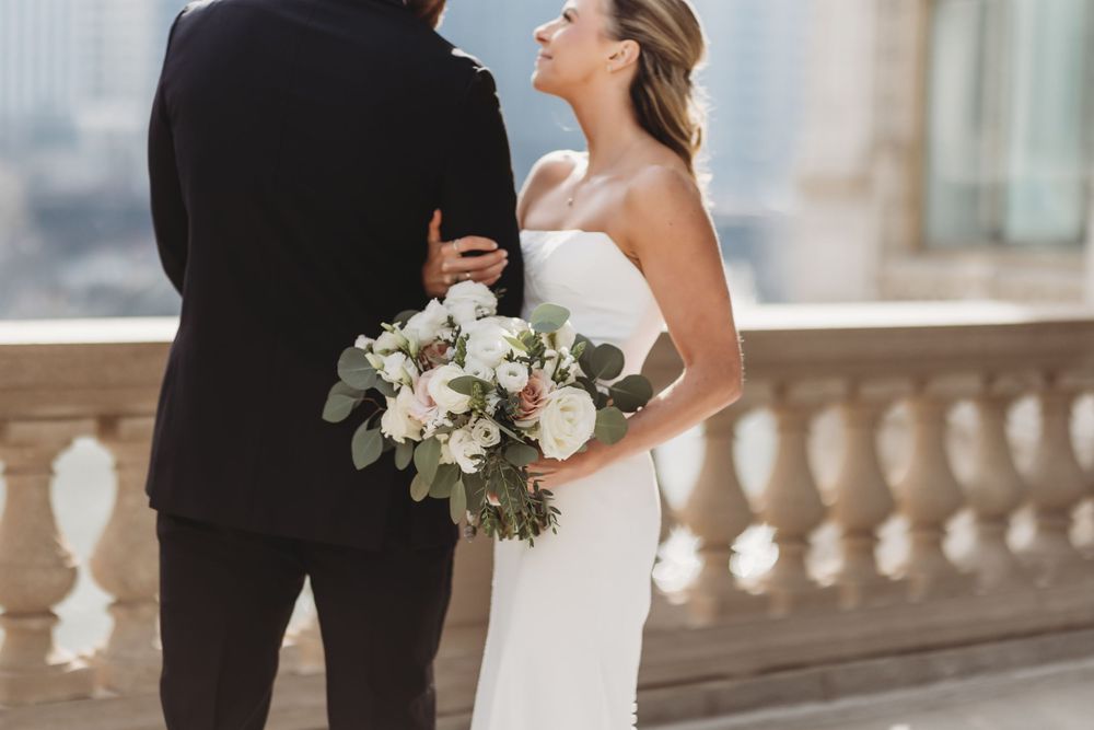 Bride in strapless white gown holds white and green bouquet beside groom in black tuxedo on terrace.