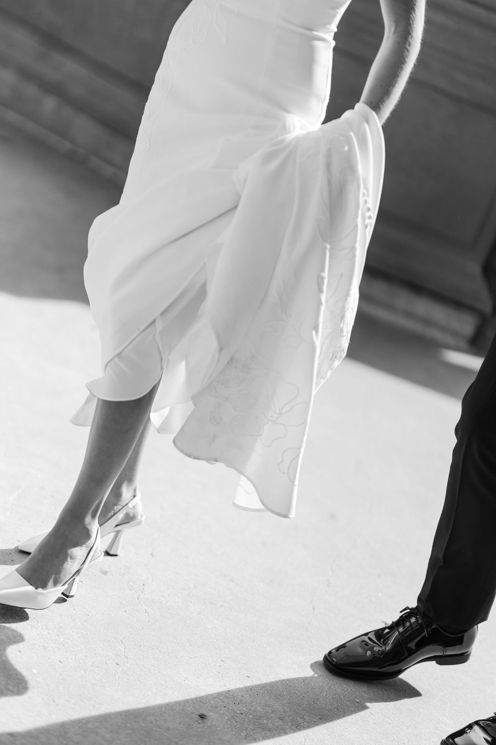 Black and white photo of bride in white gown and groom in black suit showing their wedding shoes.