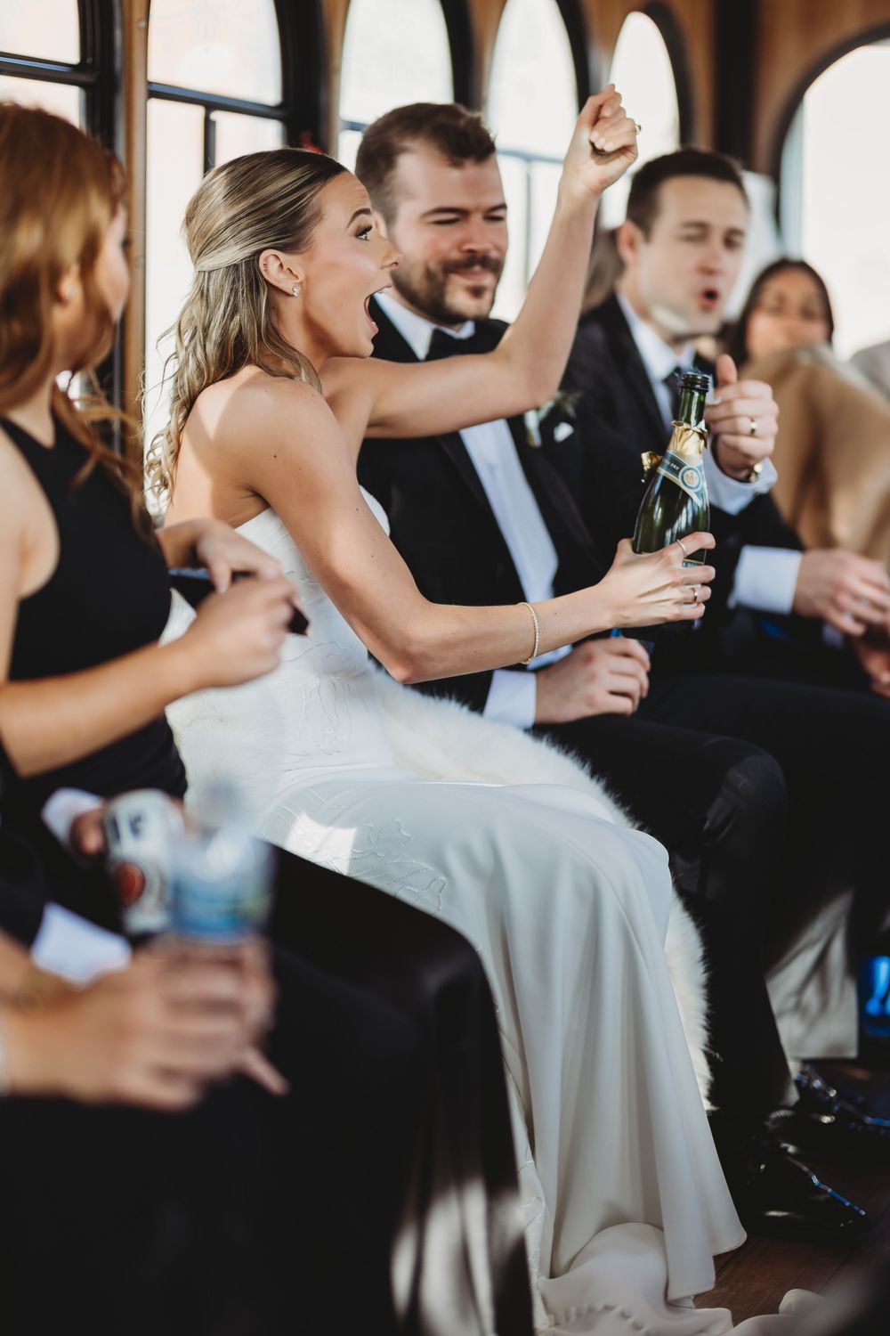 Bride in white dress pops champagne bottle on wedding party trolley surrounded by guests in formal attire.
