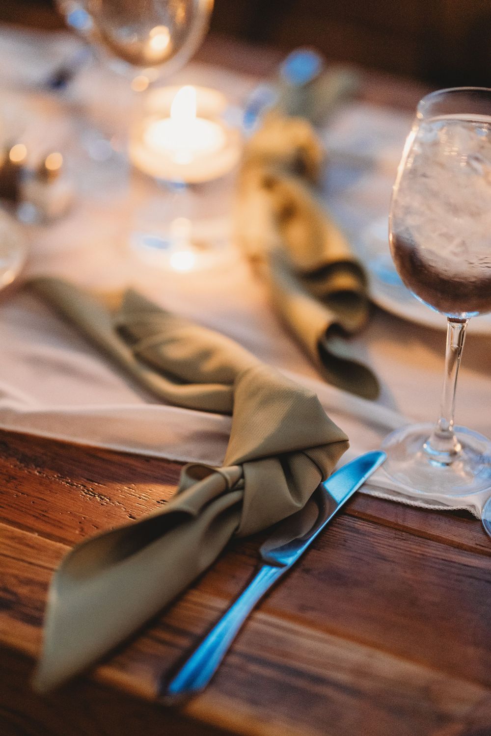 Elegant wedding table setting with sage green napkin, knife, candle, and wine glass on wood table.