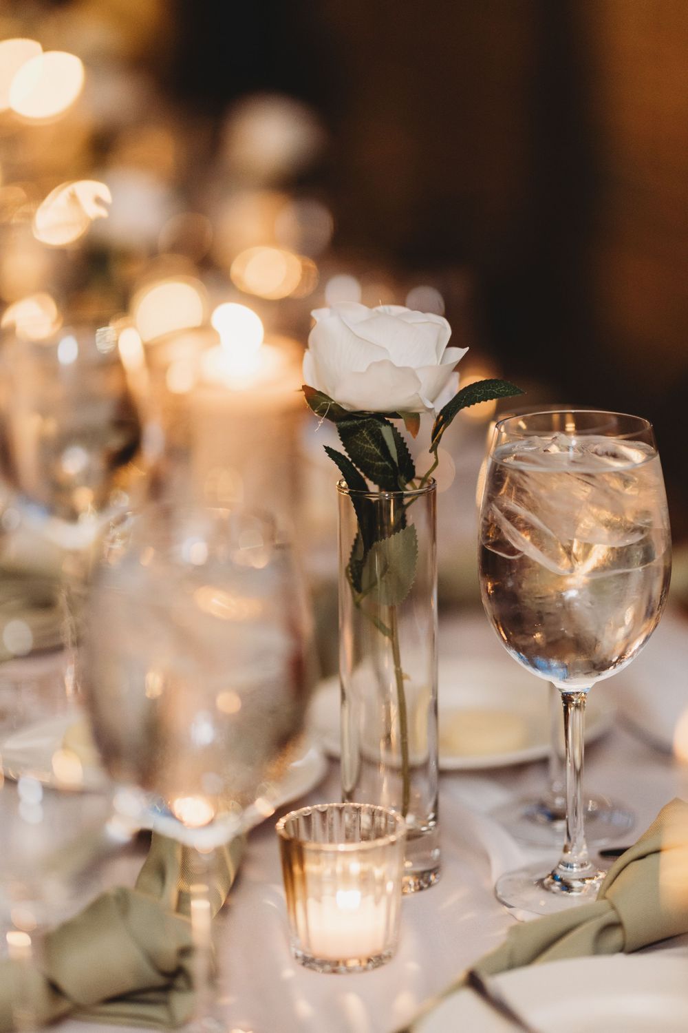 Wedding reception table with single white rose, glowing candles, and crystal wine glasses.